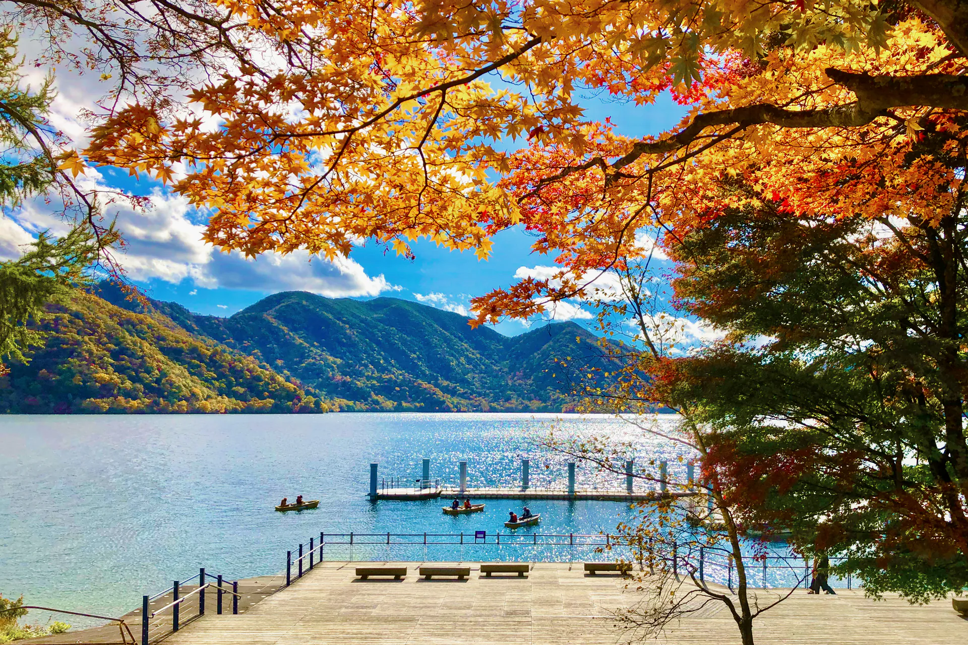 Autumn maple leaves framing Lake Chuzenji in Nikko, Japan, with rowboats by the pier and forested mountains across the water
